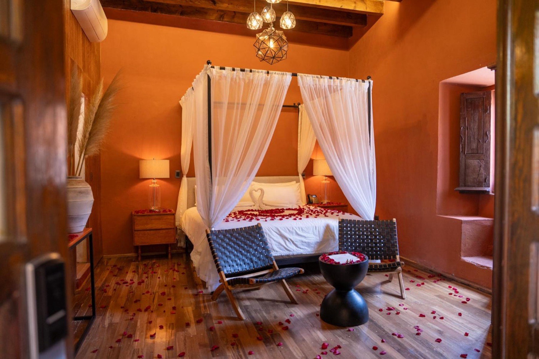Habitación de Hotel Bitos con muros en tono terracota, cama con dosel de velos blancos, vigas de madera a la vista y pétalos de rosa sobre el piso