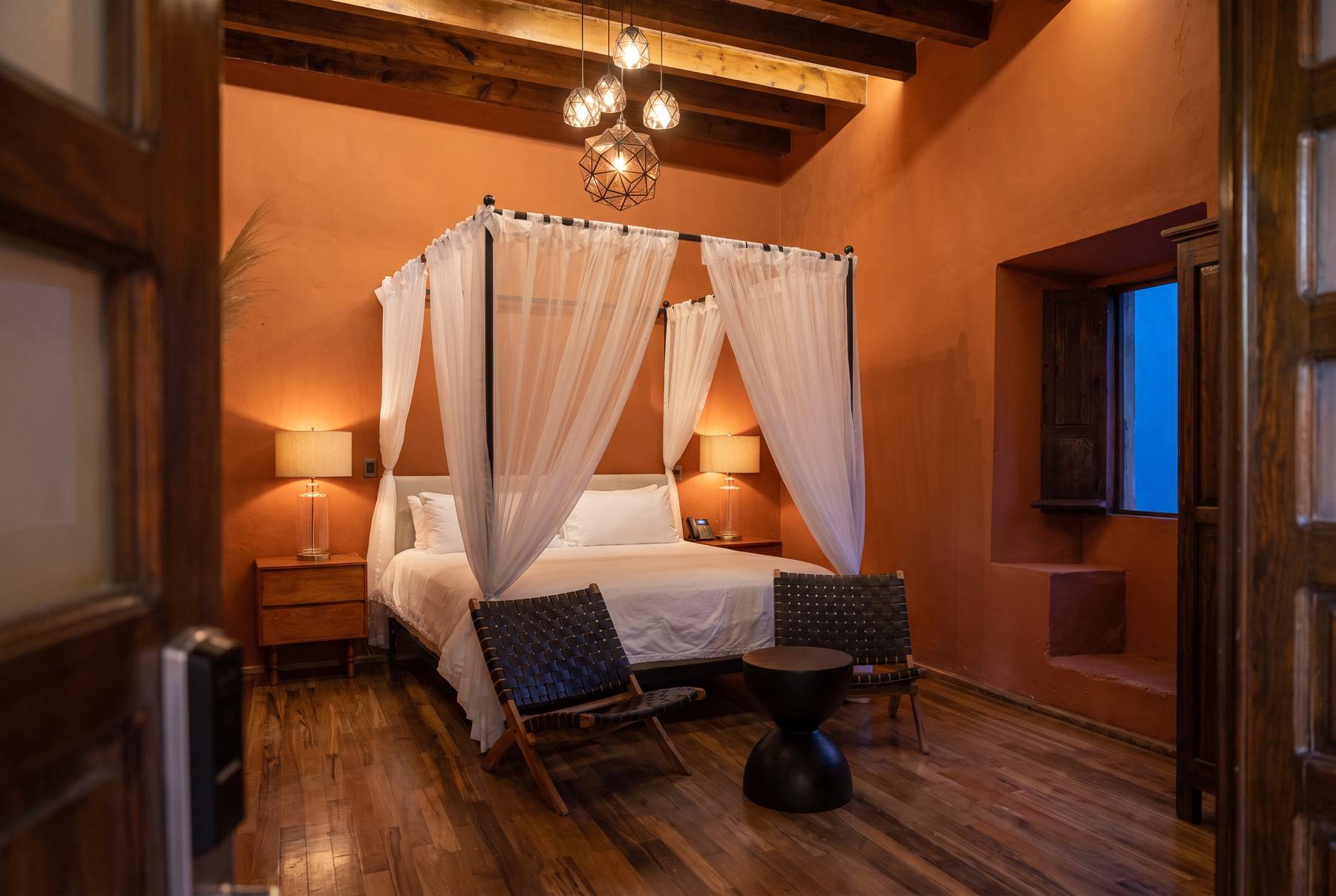 Habitación de Hotel Bitos con muros en tono terracota, cama con dosel de velos blancos, vigas de madera a la vista y piso de duela