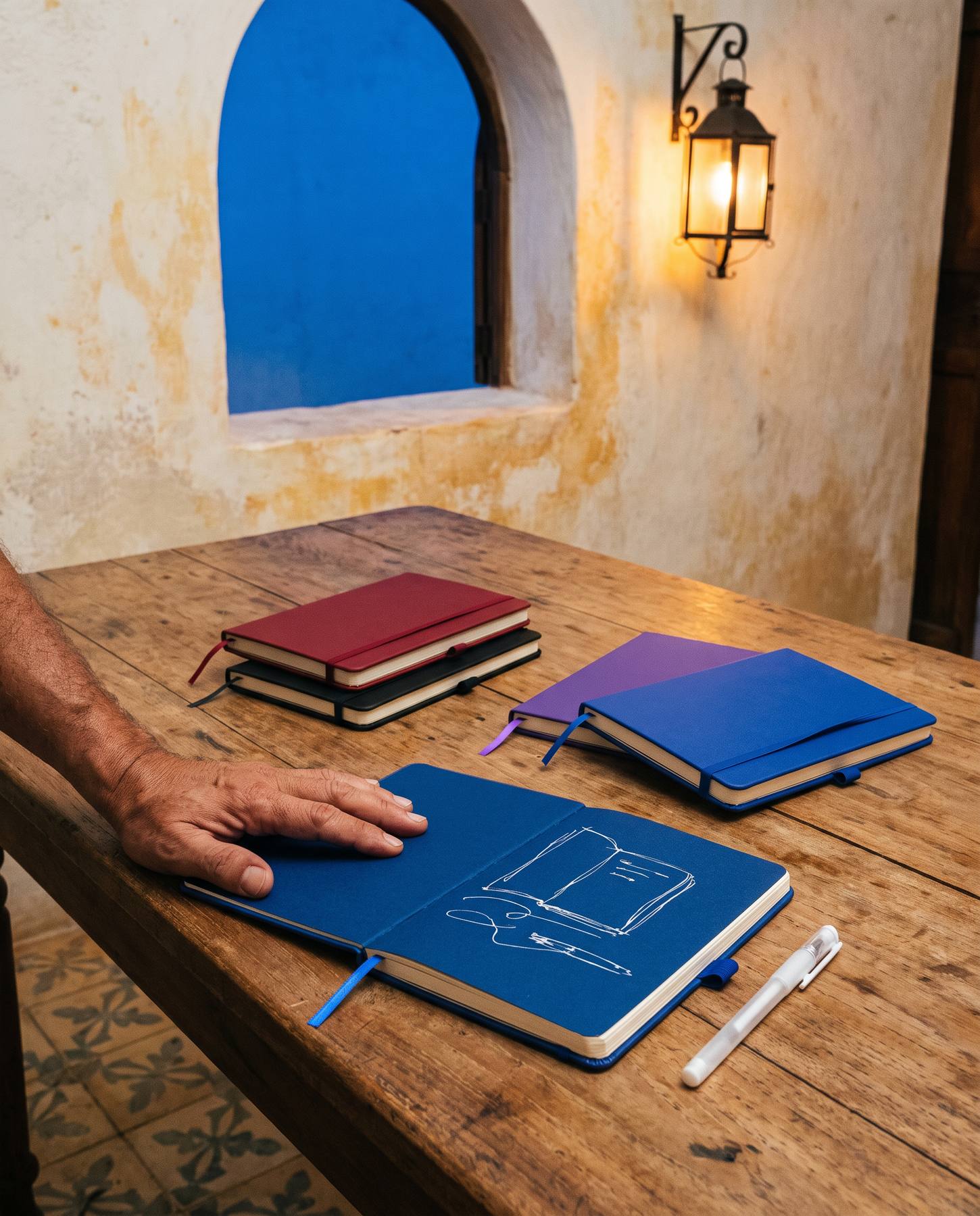 Libretas Bocetta azul, roja, morada y negra sobre una mesa de madera; una de ellas abierta, con un boceto en tinta blanca sobre la página de color