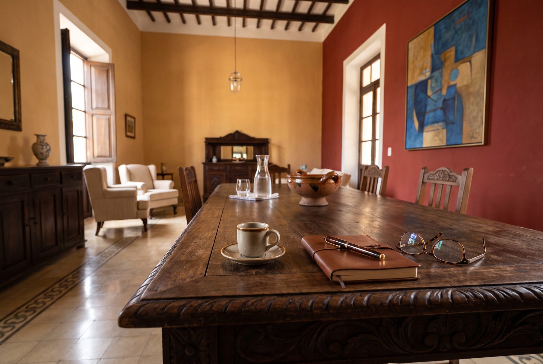 Mesa larga de una hacienda después de la comida: café, libreta de piel, pluma y lentes, con las sillas recién apartadas