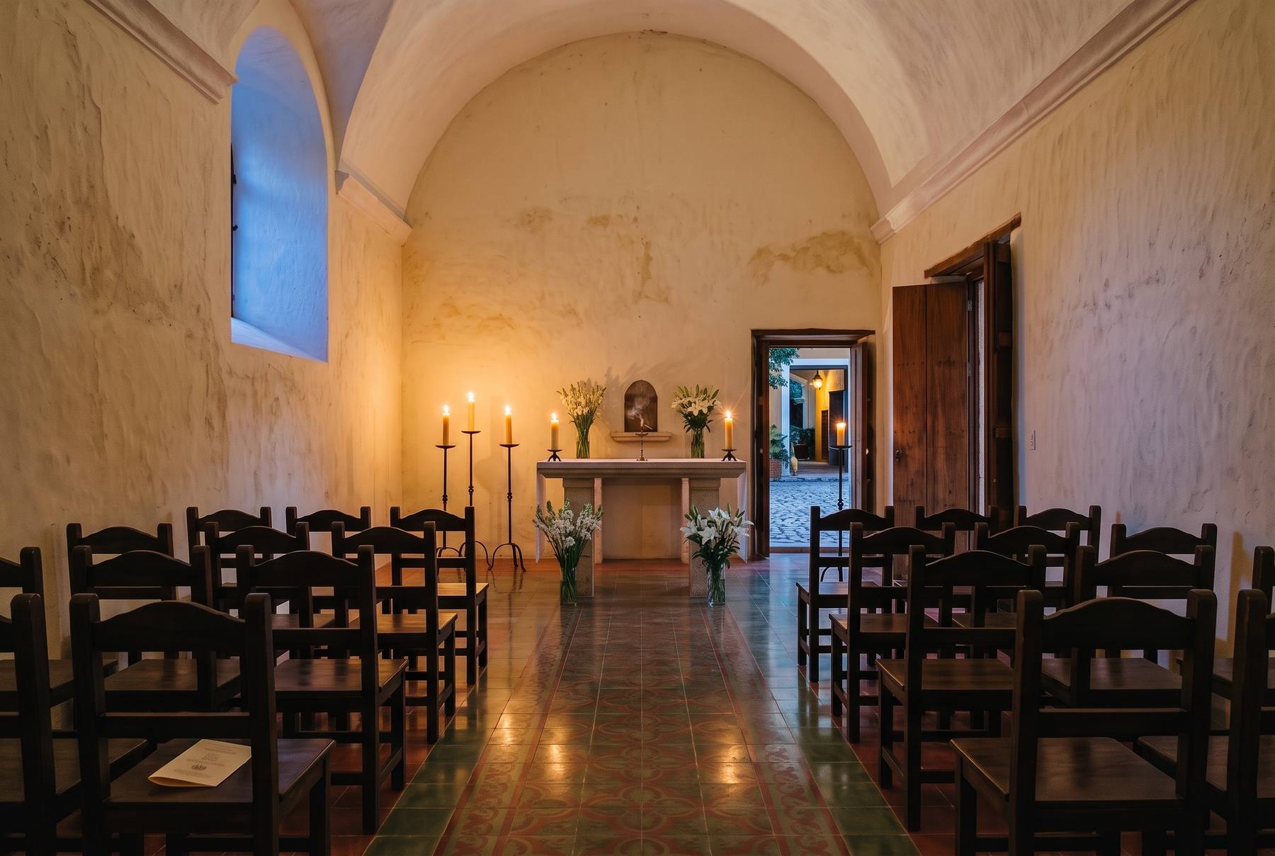 Capilla de hacienda de bóveda encalada con sillas de madera, velas encendidas y flores blancas junto al altar de piedra